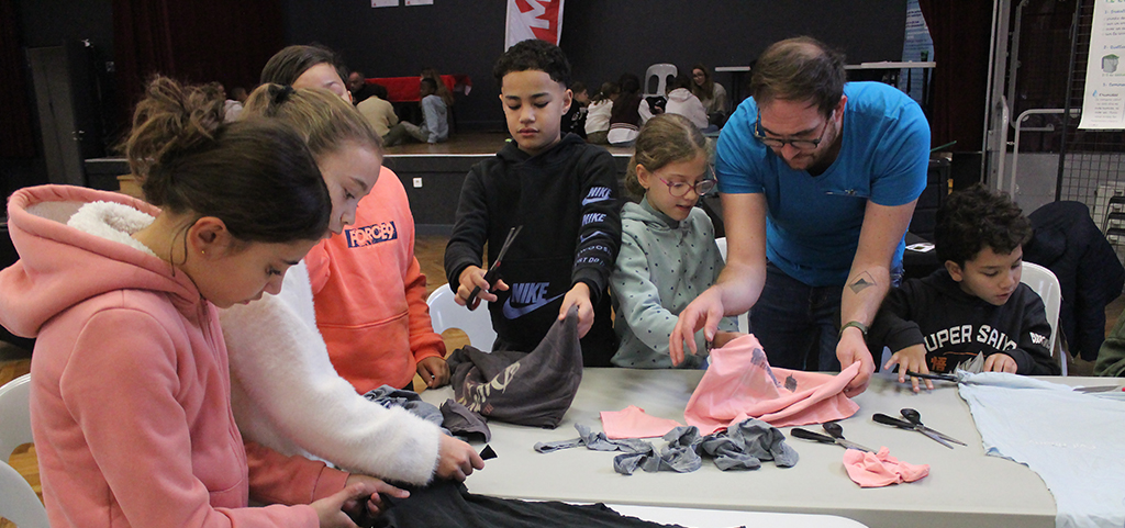 Groupe de 6 enfants et un adulte dans la salle des cordeliers, debout autour d'une table, découpent des tissus pour un atelier couture