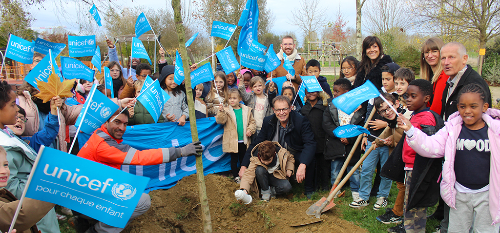 Groupe d'enfants dans le parc du couloumé à Auch, au premier plan et à gauche, agitent les drapeaux bleus de l'uncef, les élus de la ville d'Auch à droite, au centre un enfant et un adulte accroupi déposent la capsule blanc au pied de l'arbre