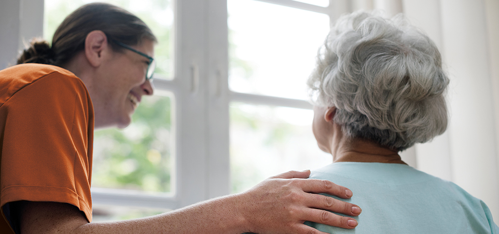 Une personne agée à droite,de dos en chemise bleu clair avec à son côté gauche son accompagnante souriante en chemise ocre, une fenêtre à l'arrière plan